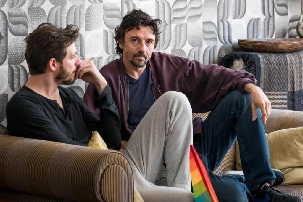 Two men sit together on a couch in a quiet, reflective scene from the French series PROUD, with a small rainbow Pride flag in the foreground.