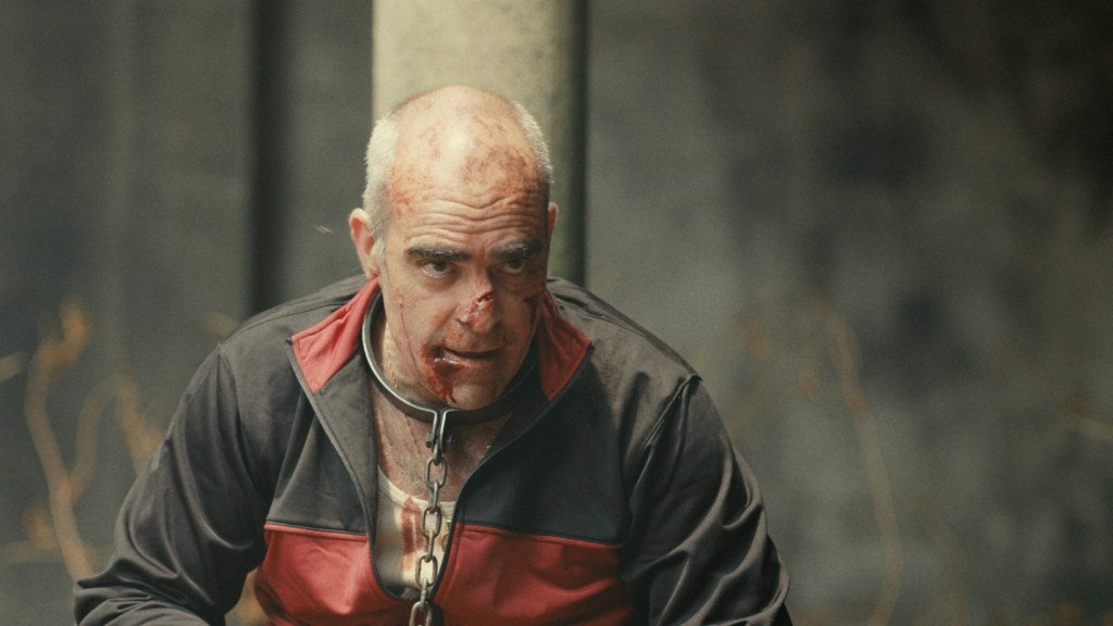 Bloodied man wearing a chain around his neck struggles to escape after being betrayed by his crime boss in a tense scene from the gay short film Concrete and Steel.