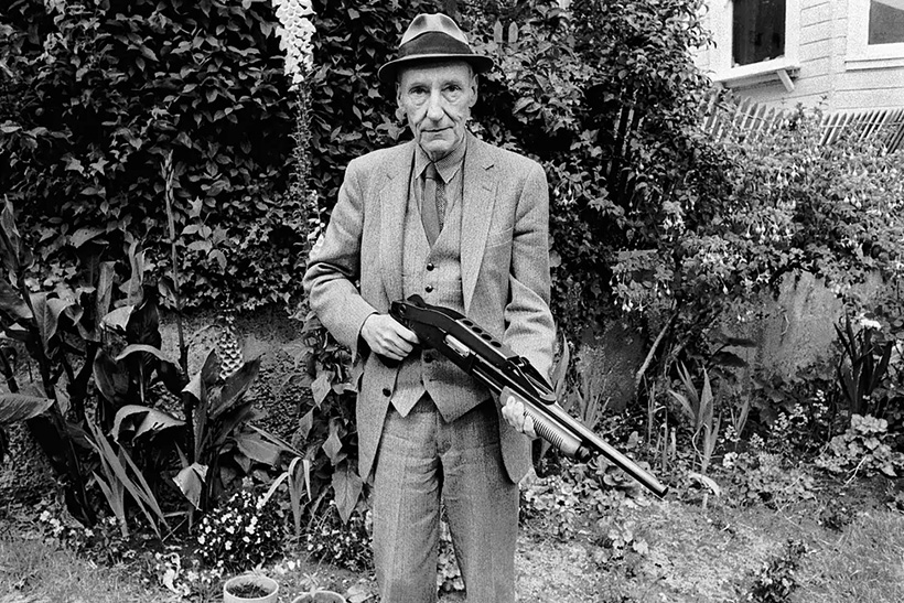 William S. Burroughs standing in a garden holding a shotgun